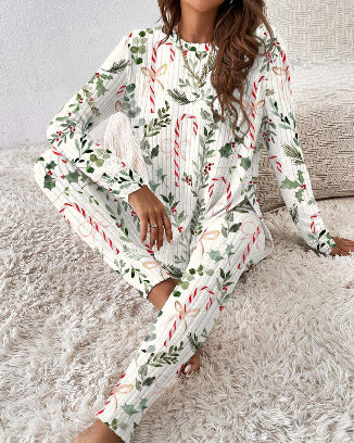 A woman wearing a long-sleeve holiday-themed pajama set with a white base and a festive print of red-and-white candy canes, green holly leaves, and pinecones, sitting on a white shag rug.